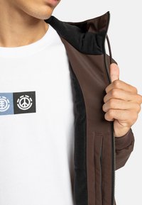 Brun lynlåsjakke med sort fleeceforet indvendig, vist delvist åben. En hvid t-shirt med trykte grafik er synlig nedenunder.