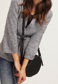 Žena v šedém pleteném kardiganu a modrých džínách, držící černou crossbody kabelku oběma rukama před sebou.