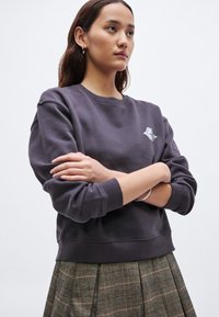 Hanorac gri închis cu guler rotund și manșete rib, având un mic logo argintiu. Asortat cu o fustă plisată în carouri verzi.