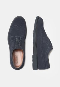 Chaussures en suede bleu foncé avec un design à lacets, présentant des détails brogue et une semelle texturée pour l'adhérence. L'intérieur affiche le nom de la marque.