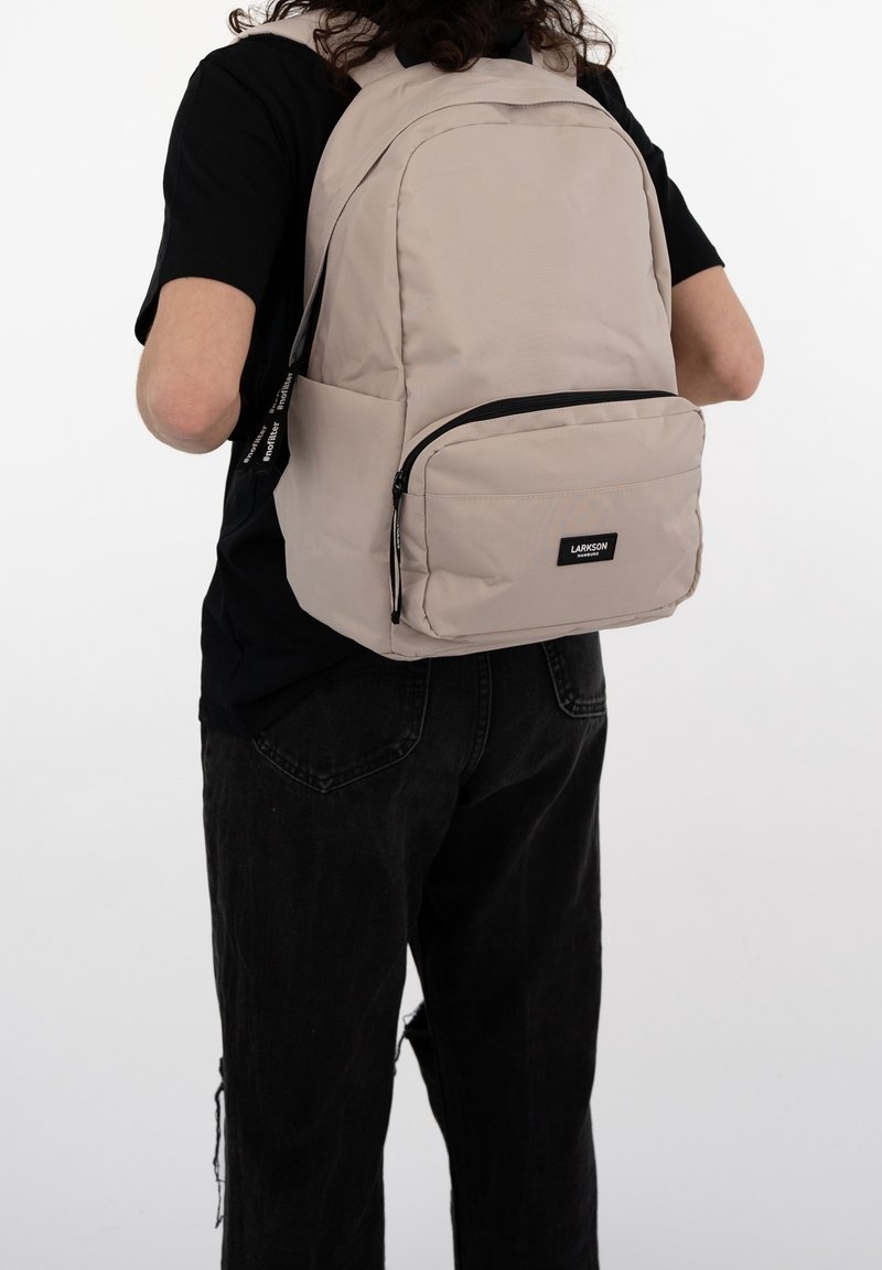 Personne portant un sac à dos beige avec une poche zippée à l'avant et une étiquette noire, habillée d'un t-shirt noir et d'un jean noir déchiré, de dos.