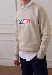 Felpa beige con logo "TOMMY JEANS" in blu e rosso, polsini a coste, indossata sopra una camicia bianca con piccoli motivi e jeans di denim scuro.