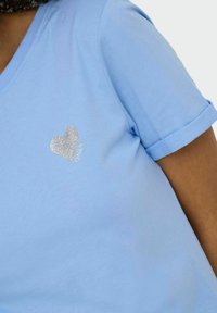 Camiseta de algodón azul claro con escote en forma de V y mangas enrolladas. Presenta un apliqué de corazón de purpurina plateada en la parte izquierda del pecho.