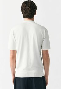 Polo majica kratkih rukava svijetlo sive boje s teksturiranim završetkom, rebrastim manžetama i rubom. Stražnja strana ima minimalistički dizajn bez vidljivih logotipa.