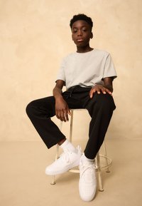 Gray cotton t-shirt, black trousers, white sneakers, and white socks. Solid colors, relaxed fit. Subject seated on a metal stool against a beige background.