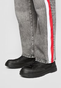 Zapatillas negras con paneles texturizados y cordones redondos, combinadas con unos jeans grises que tienen una franja lateral roja y blanca.