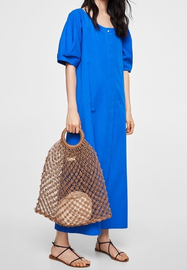 Femme portant une robe midi bleue à manches ballons, des sandales noires à brides, tenant un grand sac filet tressé avec une pochette ronde beige à l'intérieur.