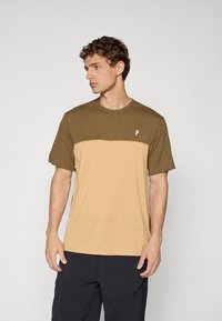 Young man with curly hair wearing a two-tone brown and beige short-sleeve t-shirt and black pants, looking left against plain background.