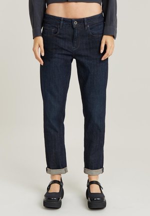 Personne portant un jean bleu foncé retroussé et des chaussures Mary Jane à plateforme noires, debout devant un fond uni.