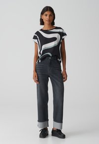 Zwarte kortemouwtop met abstracte witte en groene patronen, gecombineerd met donkergrijze opgerolde jeans en zwarte schoenen. Gladde stof en relaxte pasvorm.