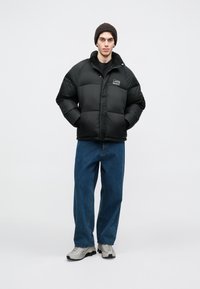 Μαύρο μπουφάν τύπου πούφες με φαρδιά εφαρμογή, που διαθέτει ραμμένα τετράγωνα κοψίματα και λογότυπο στο στήθος. Φοράει ανοιχτόχρωμες τζιν παντελόνες με φαρδιά γραμμή και γκρίζα αθλητικά παπούτσια.