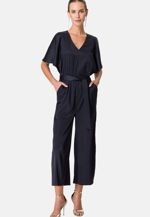 Combinaison marine en tissu lisse avec un décolleté en V, manches courtes, taille cintrée, pantalons à jambes larges et poches latérales. Assortie à des talons dorés.