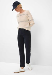 Pull beige rayé avec une texture douce, pantalon noir ajusté et baskets blanches. La casquette est bleu marine, ajoutant un contraste à la tenue.