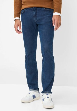 Jean en denim bleu foncé avec une coupe droite, présentant un design classique à cinq poches, associé à des baskets blanches et marines.