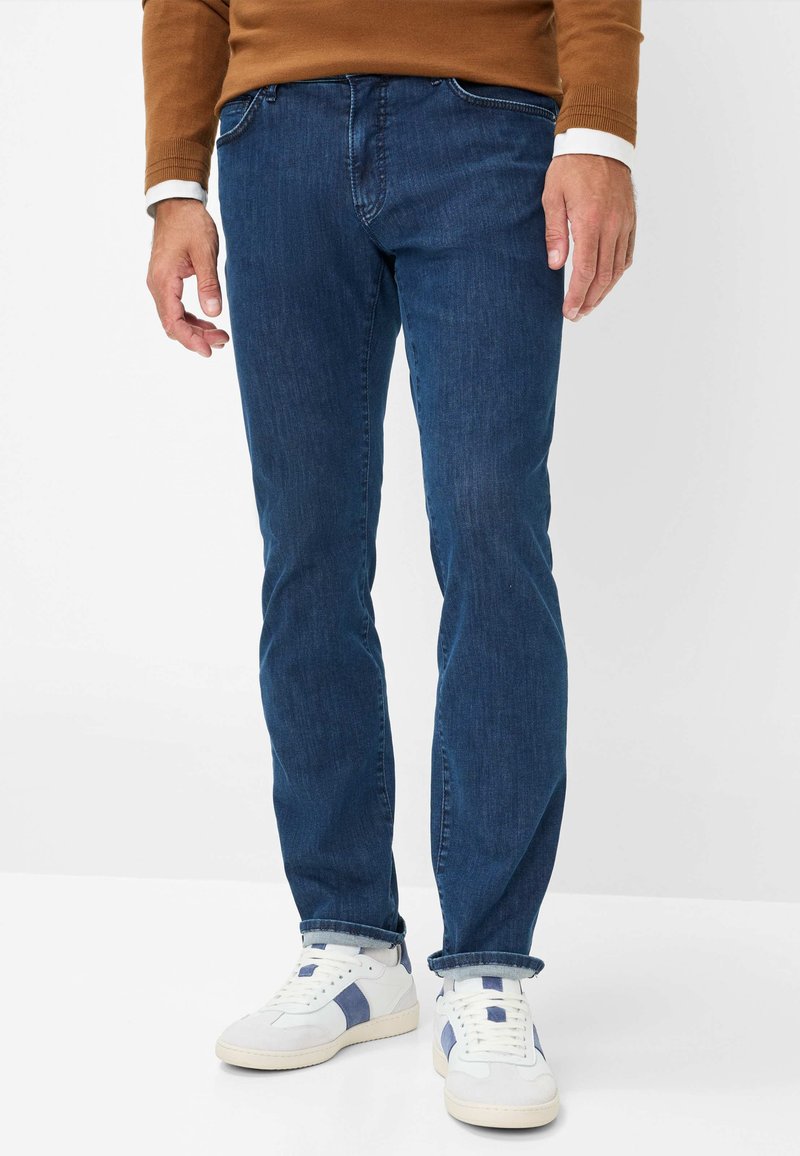 Jeans in denim blu scuro con vestibilità dritta, caratterizzati da un classico design a cinque tasche, abbinati a sneaker bianche e blu navy.