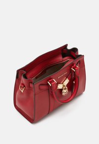 Sac à main en cuir rouge avec une forme structurée, doubles poignées supérieures, quincaillerie dorée et charm pendentif avec logo. L'intérieur comporte une poche zippée.