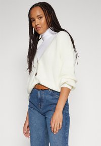 Cream-colored knitted cardigan with a V-neck, featuring two buttons and loose sleeves. Paired with high-waisted blue denim jeans and a brown belt.