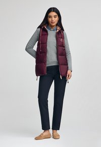 Bordeauxfarbene Weste mit Steppdesign, ausgestattet mit einer braunen satinbeschichteten Kapuze und goldenen Beschlägen. Kombiniert mit einem grauen Pullover und dunklen Hosen.