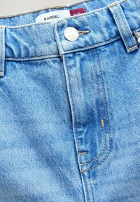Jeans din denim de un albastru deschis cu cusături portocalii, având o închidere cu buton, un eticheta marcata "BARREL" și un aspect texturat al materialului.
