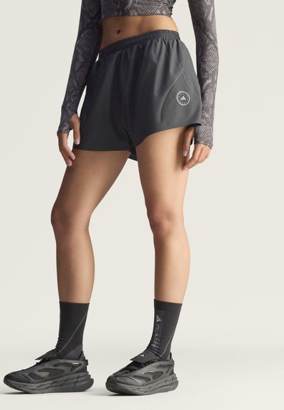 Person trägt schwarze Laufsportshorts, schwarze Sportschuhe, schwarze Socken mit weißem Schriftzug und ein langärmeliges, gemustertes Crop-Top und steht.