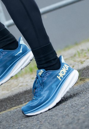 Blå Hoka løbesko båret med sorte leggings på en udendørs asfaltflade nær græs og kantsten.
