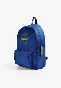 Mochila de nailon azul con la parte superior redondeada, bolsillo frontal con cremallera y logo verde. Cuenta con correas ajustables negras para los hombros y un tirador de cremallera texturizado.