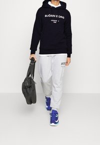 Felpa con cappuccio blu navy con testo bianco, pantaloni della tuta grigi e scarpe blu con accenti verdi. Borsa nera con tracolla.