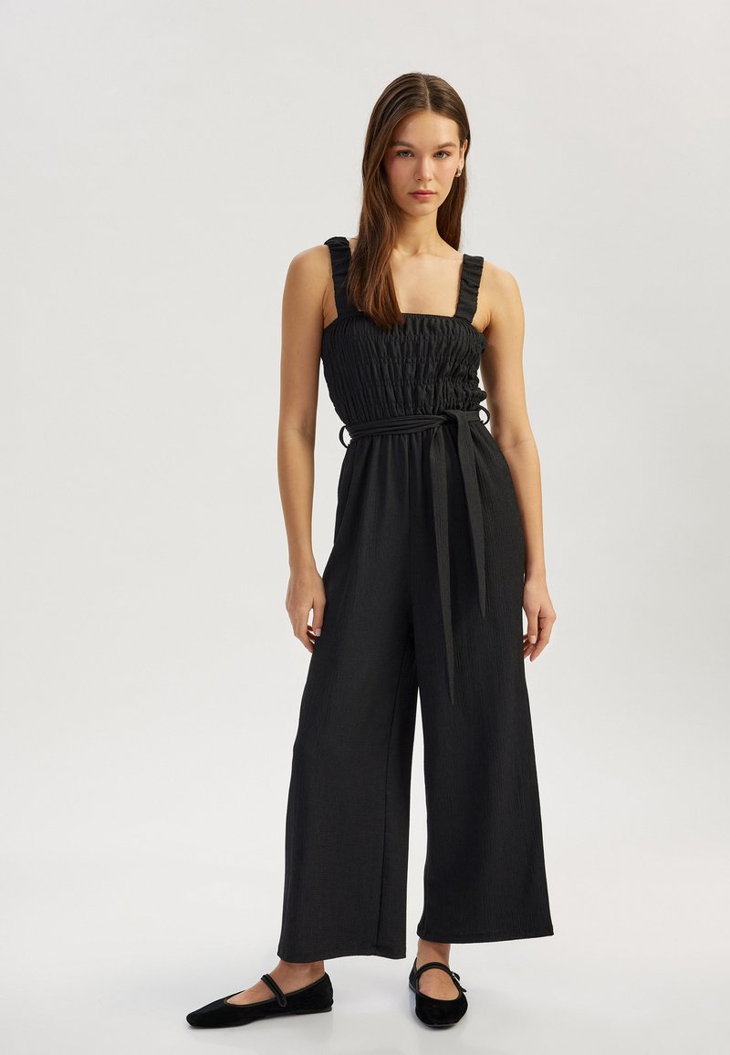 Mono de pierna ancha negro con cuerpo fruncido, tirantes con volantes y un cinturón a juego. La suave tela texturizada complementa una silueta fluida.