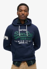 Felpa con cappuccio blu navy con grafiche verdi e bianche. Presenta una tasca a marsupio e una camicia a quadri in contrasto sotto. Design casual.