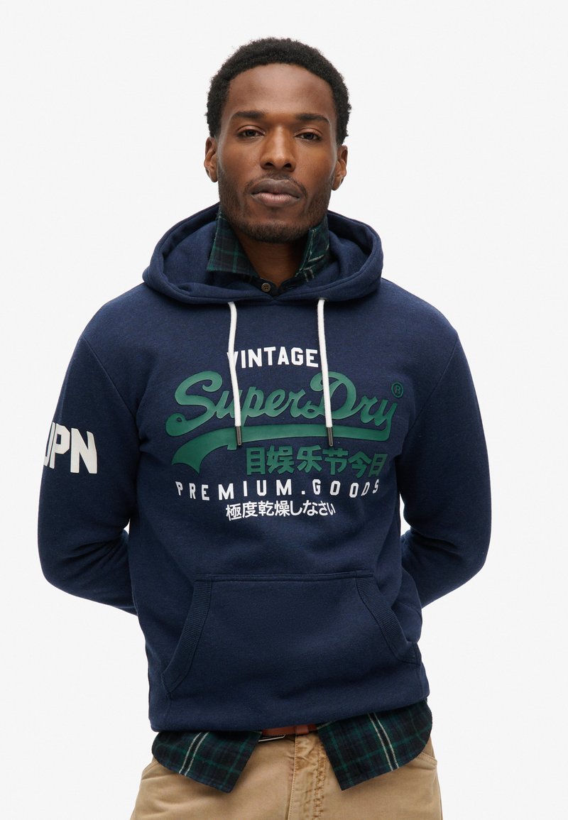 Felpa con cappuccio blu navy con grafiche verdi e bianche. Presenta una tasca a marsupio e una camicia a quadri in contrasto sotto. Design casual.