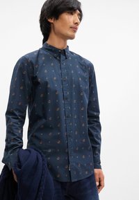 Μπλε πουκάμισο με κουμπιά και λουλουδάτο μοτίβο, φτιαγμένο από μαλακό ύφασμα. Οι μακριές μανίκες και ο γιακάς με κουμπιά συμπληρώνουν το σχέδιο.