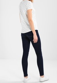 Camiseta blanca de manga corta con estampado de ancla negro, combinada con pantalones ajustados de mezclilla azul oscuro y zapatillas blancas. Vista desde la parte de atrás.