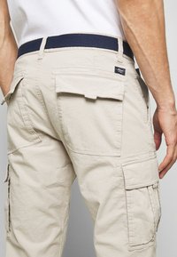 Pantalones cargo beige con múltiples bolsillos laterales, tejido texturado y una cinturilla elástica azul marino. Presenta acentos cosidos y una pequeña etiqueta.