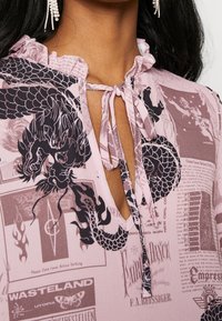 Růžová blůza s volánkovým výstřihem, předním zavazováním a černým potiskem draka. Doprovodný text a vzory vytvářejí texturovaný design.