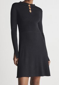 Robe en tricot côtelé noire à manches longues, dotée de trois boutons décoratifs au niveau du col et d'entailles sur les côtés de l'ourlet pour plus de détails.