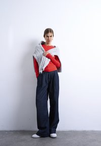 Γυναίκα που στέκεται μπροστά σε λευκό τοίχο φορώντας κόκκινη μπλούζα με μακριά μανίκια, σκούρο παντελόνι με φαρδιά πόδια, λευκά παπούτσια και γκρι πλεκτή ζακέτα ριγμένη στους ώμους της.