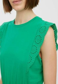 Zelené bavlněné tričko s krajkovými detaily na ramenou a květinovými výstřihy. Kulatý výstřih a volný střih.
