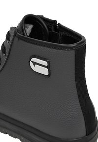 Tuvplāns melna ādas potītes zābaka ar teksturētu virsmu, melnām šņorēm, polsterētu apkakli un sudraba un melna logotipa uz sāniem.