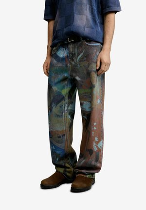 Jeans larghi con stampe artistiche multicolori di figure umane, indossati con scarpe marroni e una camicia a quadri blu scuro.