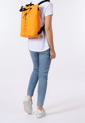 Zaino impermeabile arancione con chiusura a rotolo, spallacci neri e logo grande. Texture liscia, forma rettangolare e senza motivi visibili.