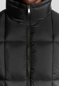 Schwarze Pufferjacke mit hohem Kragen, durchgehendem Reißverschluss und gestepptem Designs, das eine glatte, glänzende Textur aufweist.