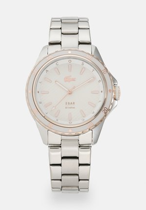 Reloj de acero inoxidable plateado con bisel de oro rosado, esfera texturizada blanca y marcadores luminosos. Presenta una pulsera de eslabones y una corona para ajustes.