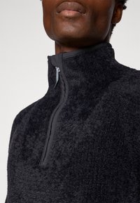 Svart fleece-pullover med hög krage, som har en kvartzip-stängning. Mjuk textur och en kontrasterande svart kant runt halsen.