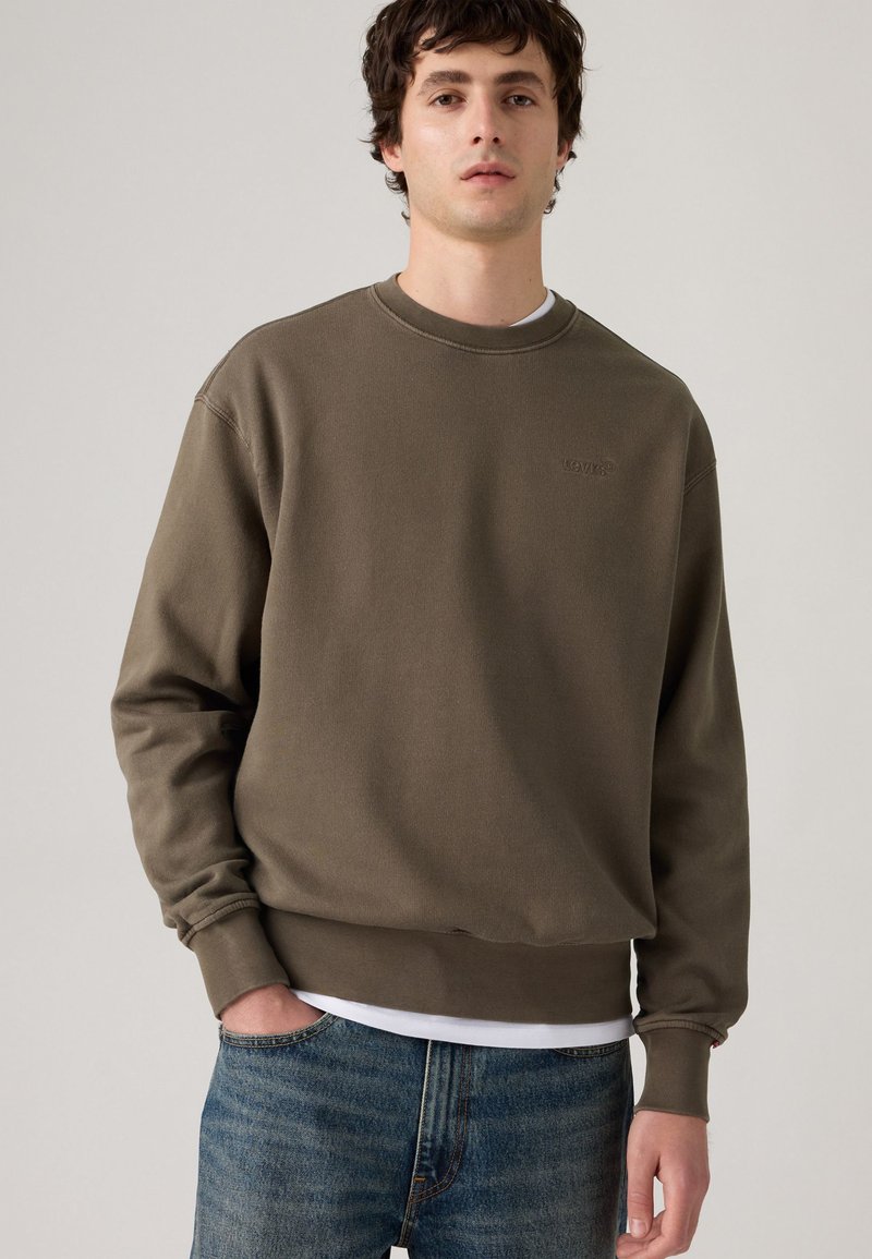Sudadera verde oliva de corte relajado, con cuello redondo y puños acanalados, que presenta un pequeño logo en el pecho. Llevada sobre una camiseta blanca.