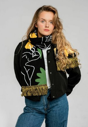 Czarny szalik z żółtymi i zielonymi wzorami, z białymi konturami i złotą frędzlem. Model nosi czarną koszulę i niebieskie dżinsy.