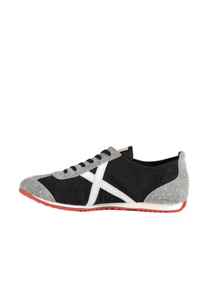 Zapatilla deportiva negra y gris con rayas laterales blancas, cordones negros y suela roja, mostrada desde la vista lateral exterior sobre un fondo blanco.
