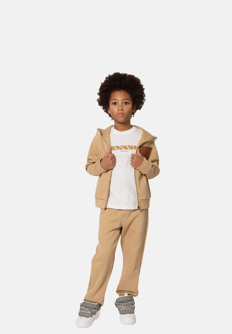 Tan zip-up hoodie en bijpassende broek van zachte stof, gecombineerd met een wit t-shirt met bruin grafisch opdruk. Schoenen hebben zwart-witte accenten.