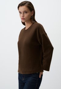 Maglione marrone a maniche lunghe con una finitura testurizzata, scollatura rotonda e spalle abbassate, abbinato a jeans blu scuro.