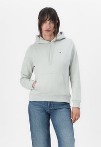 Hellgrüner Hoodie aus weichem Material mit Kordelzügen, einer Fronttasche, gerippten Bündchen und einem kleinen Logo auf der Brust. Kombiniert mit blauen Jeans.