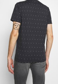 Tmavě šedé tričko s krátkým rukávem, bílé geometrické vzory, kulatý výstřih a měkká bavlněná textilie. Pohled zezadu ukazuje délku a střih trička.